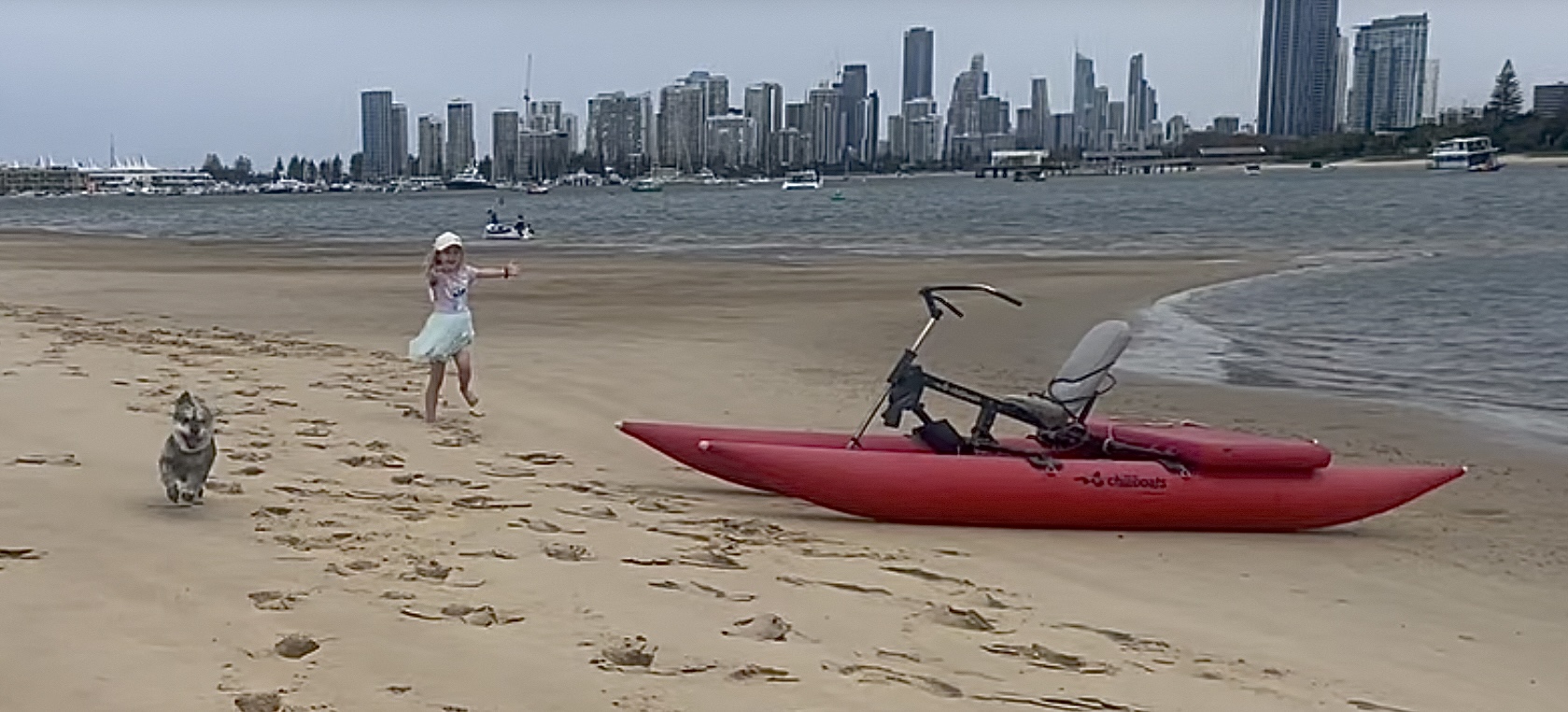 A red ChiliBoats waterbike on the beach with a child and dog, overlooking the Gold Coast skyline